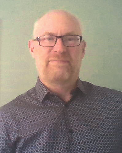 Stuart Hamby smiling with folded arms, leaning against historic stone wall